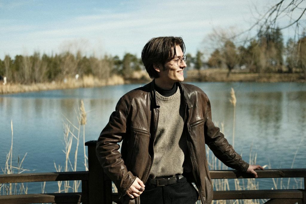 A man in a brown jacket and glasses standing by a lake